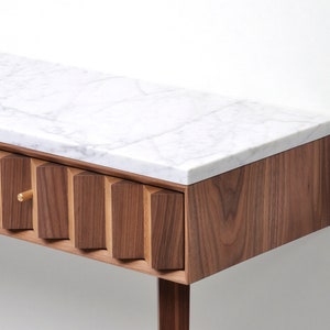 May include: A modern walnut wood console table with a white marble top and a single drawer. The drawer front features a unique geometric design with a brass knob.