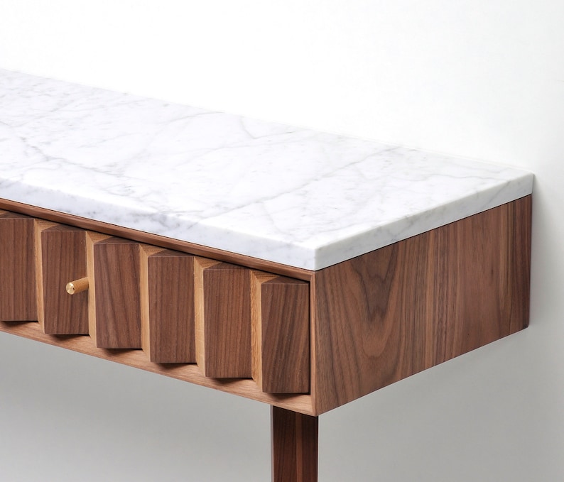 May include: A modern walnut wood console table with a white marble top and a single drawer. The drawer front features a unique geometric design with a brass knob.