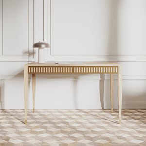 Puede incluir: Un escritorio de madera clara con detalles dorados y dos cajones. El escritorio tiene un diseño sencillo con patas cónicas y una tapa rectangular. El escritorio está sobre un suelo con estampado.