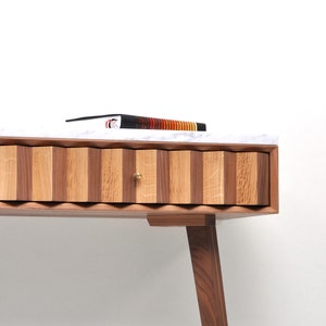 May include: A wooden desk with a white marble top and a single drawer. The drawer has a wavy, scalloped front and a brass knob. The desk has two walnut legs.
