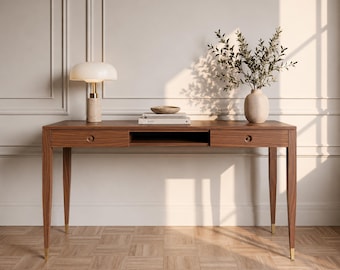 May include: A rectangular wooden desk with two drawers featuring circular pulls, a central open storage compartment, and four tapered legs with gold-toned metal feet. A mushroom-shaped lamp, a stack of books, and a vase with greenery sit on top.