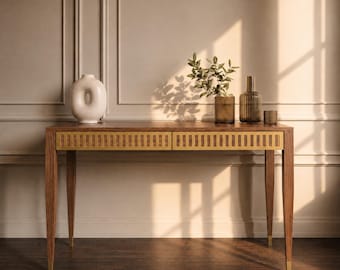 May include: A wooden console table with two drawers featuring vertical brass slats on the front. The table has four tapered legs with brass feet. On top sits a white donut-shaped vase, a glass vase with green leaves, and two glass containers.