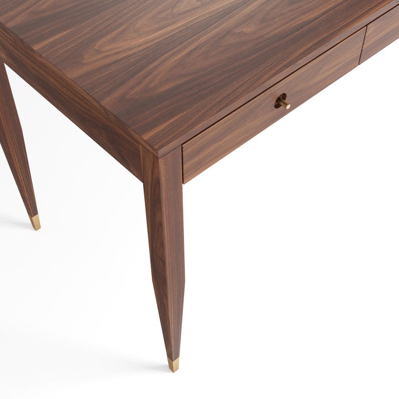 May include: A dark brown walnut wood desk featuring a rectangular top with visible wood grain patterns, a single drawer with a small cylindrical brass knob, and four tapered legs that each terminate in a polished brass metal foot cap.