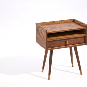 May include: A wooden bedside table with a drawer and a shelf. The table has four tapered legs with gold accents.