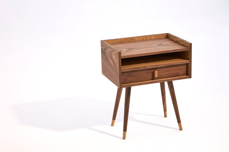 May include: A wooden bedside table with a drawer and a shelf. The table has four tapered legs with gold accents.