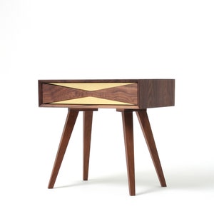 May include: A walnut wood nightstand with a single drawer featuring a brass inlay in a geometric pattern. The nightstand has four legs.