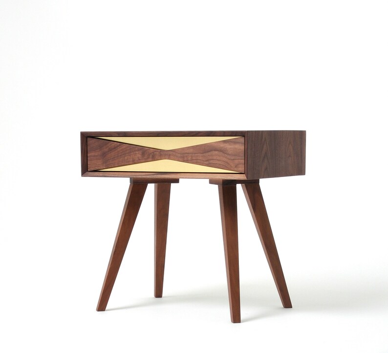 May include: A walnut wood nightstand with a single drawer featuring a brass inlay in a geometric pattern. The nightstand has four legs.