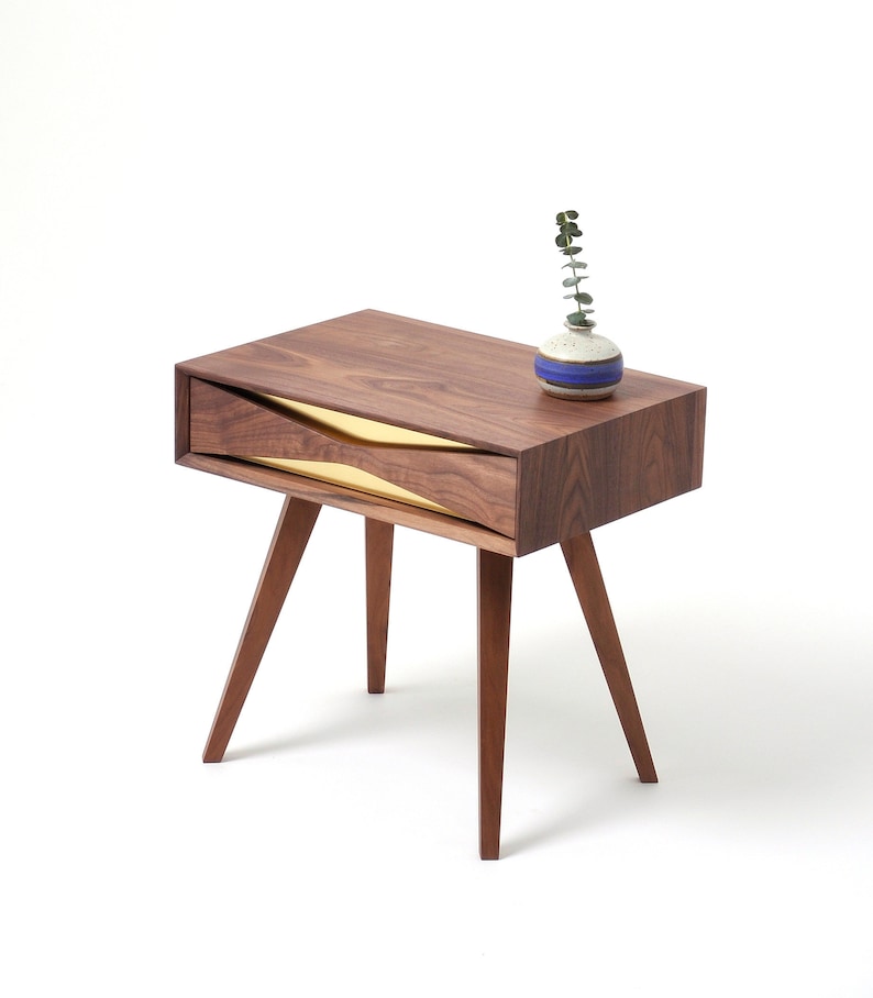 May include: A wooden end table with a single drawer featuring a gold-colored interior. The table has four legs and a small vase with a sprig of greenery on top.