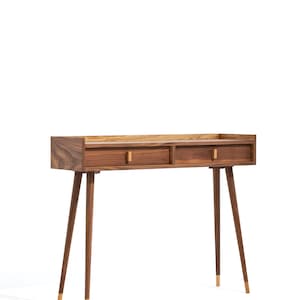 Puede incluir: Una mesa consola de madera con dos cajones y patas cónicas. La mesa está hecha de madera de nogal y tiene un acabado natural. Las patas están acabadas con detalles dorados.