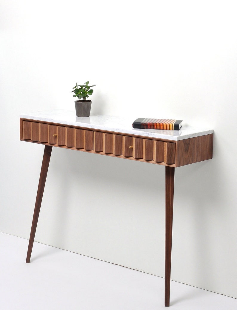 May include: A walnut wood console table with a white marble top and two drawers. The table has a fluted design on the front and tapered legs.