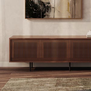 Op de afbeelding: Een houten tv-meubel in mid-century modern stijl, met een lamellen voorkant. De console heeft een warme bruine kleur en zwarte metalen poten. De achtergrond toont een abstract schilderij en een neutraal gekleurde muur.