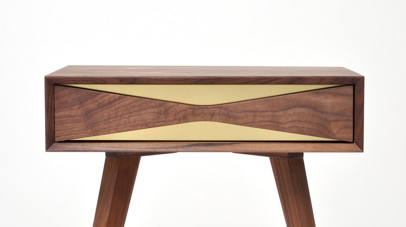 May include: A walnut wood nightstand with a single drawer featuring a brass inlay. The drawer has a unique, angled design. The nightstand has four legs.