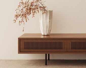 May include: A modern wooden media console with a slatted front design, supported by black metal legs. A textured white vase holds dried branches with small, brown leaves. The console is a warm brown color, and the background is a neutral beige.