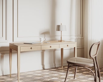 Op de afbeelding: Een licht houten bureau met drie laden en een lamp erop. Een gevlochten stoel staat voor het bureau. Het bureau en de stoel bevinden zich in een kamer met witte muren en een betegelde vloer.