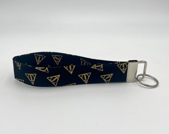 May include: Black key chain with a gold Deathly Hallows symbol pattern. The key chain has a silver metal ring.