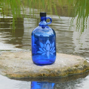 Könnte beinhalten: Eine blaue Glasflasche mit einem weißen Lotusblüten-Design steht auf einem Stein in einem Teich. Die Flasche hat einen Henkel und einen schwarzen Deckel.