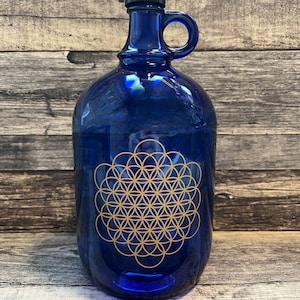 Op de afbeelding: Een blauwe glazen fles met een gouden geometrisch ontwerp. De fles heeft een handvat en een zwarte dop.