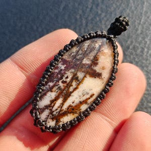 Puede incluir: Un colgante de forma ovalada que presenta una piedra pulida con vetas blancas, marrones y moradas oscuras. La piedra está encerrada en un borde de macramé negro hecho de cordón anudado, con un pequeño lazo en la parte superior para colgar.