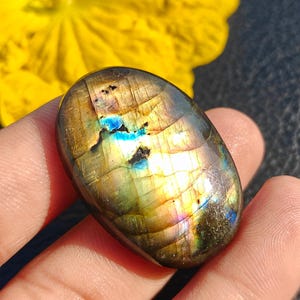 May include: An oval-shaped labradorite gemstone featuring a polished surface with iridescent flashes of gold, blue, and green light. The stone displays natural internal fractures and dark inclusions within its smooth, rounded, and reflective structure.