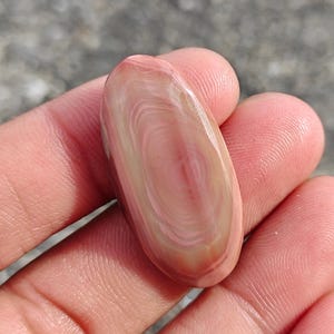 Op de afbeelding: Een gepolijste, ovaalvormige edelsteen met een wervelend patroon van roze, crème en lichtbruine tinten. De steen, vastgehouden in een hand, heeft een glad, glanzend oppervlak en natuurlijke kleurvariaties. De edelsteen is ongeveer 3,8 cm lang.
