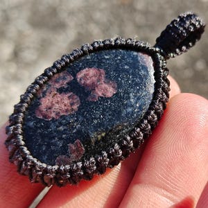Peut inclure: Un pendentif en pierre bleu foncé et marron avec un cordon macramé. La pierre présente des taches marron et est entourée d'une bordure macramé noire. Le pendentif est tenu dans une main.
