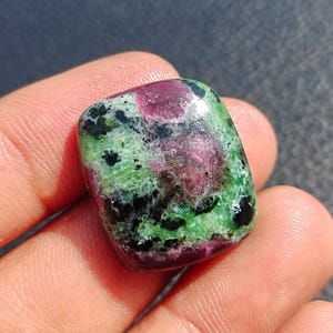 Peut inclure: Une pierre précieuse rectangulaire polie avec un mélange de couleurs. La pierre présente un centre rouge profond, entouré d'inclusions vertes et noires. La pierre est tenue dans une main, mettant en valeur sa beauté naturelle.