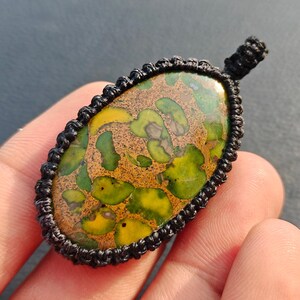 May include: An oval-shaped pendant featuring a polished stone with a mottled pattern of vibrant green, yellow, and brown earthy tones. The stone is securely encased in a black cord frame created with intricate macrame knotting techniques.