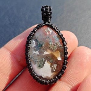May include: An oval-shaped pendant featuring a polished moss agate stone with translucent white, brown, and green dendritic inclusions, encased in a black macrame cord setting with a matching woven loop at the top for suspension.