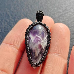 May include: A teardrop-shaped amethyst pendant featuring a polished purple and white chevron-patterned stone encased in a black macrame cord setting with a small knotted loop at the top for hanging on a necklace chain or cord.