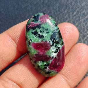 May include: An oval gemstone displaying a mix of colours. The stone has deep red, green, and black hues. The red areas are in patches, while the green and black create a speckled pattern. The gemstone is held in a hand.