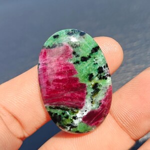 Puede incluir: Una piedra preciosa en cabujón de forma ovalada que presenta una mezcla vibrante de inclusiones de rubí rojo magenta intenso sobre una matriz de zoisita verde moteada con motas negras de hornblenda, pulida con un acabado liso.