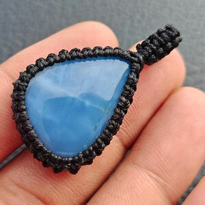 May include: A teardrop-shaped blue stone pendant encased in a black macrame cord setting. The smooth, polished blue gemstone features a vibrant, solid color and is framed by intricate, knotted black cord work that forms a loop at the top.