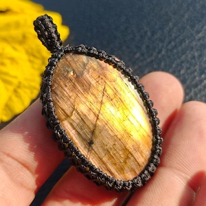 May include: An oval-shaped labradorite gemstone pendant featuring a polished, iridescent surface with golden and brown tones, encased in a dark black macrame cord setting with a matching woven loop at the top for hanging as a necklace piece.