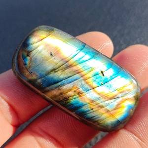 May include: A rectangular labradorite gemstone featuring a polished surface with iridescent flashes of blue, gold, and yellow. The stone displays natural internal fractures and a smooth, rounded edge profile, showcasing a vibrant play of colour.