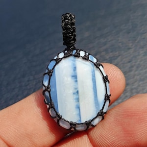 May include: An oval-shaped pendant featuring a smooth, polished stone with alternating vertical stripes of white and light blue. The stone is secured within a black cord macrame setting that forms a decorative net pattern around the edges.
