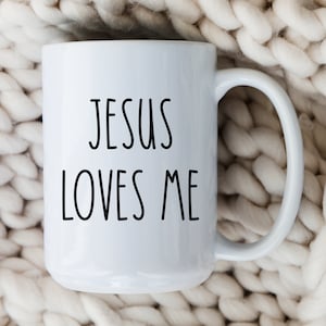 May include: White ceramic mug with black text that reads "Jesus Loves Me". The mug is sitting on a white knitted blanket.