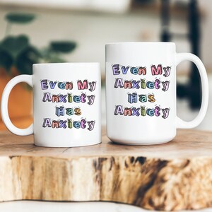 May include: Two white coffee mugs with the text "Even My Anxiety Has Anxiety" in colorful, outlined letters. The mugs are on a wooden surface.