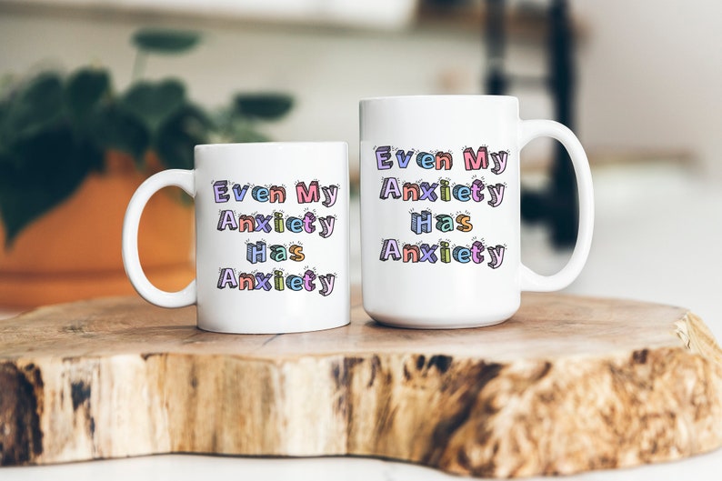 May include: Two white coffee mugs with the text "Even My Anxiety Has Anxiety" in colorful, outlined letters. The mugs are on a wooden surface.