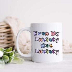 May include: White ceramic coffee mug with a colorful design that reads "Even My Anxiety Has Anxiety".