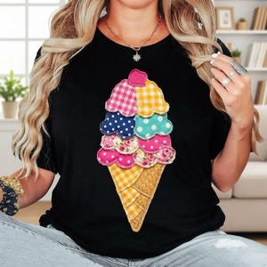 May include: Black t-shirt featuring a colorful ice cream cone design. The ice cream has layers of pink, blue, and yellow scoops with various patterns, including polka dots and floral prints. The cone is a yellow and brown waffle pattern.