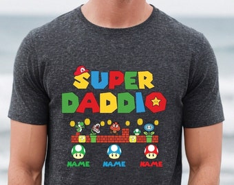 May include: A dark gray t-shirt with the text "SUPER DADDIO" in colorful block letters. The text is styled to look like a video game level with a star and mushrooms.