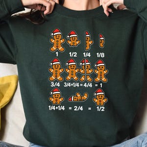 May include: Dark green sweatshirt featuring gingerbread men wearing Santa hats. The sweatshirt displays fractions with gingerbread men, including 1, 1/2, 1/4, and 1/8. Equations such as 3/4 + 1/4 = 4/4 = 1 are also shown.