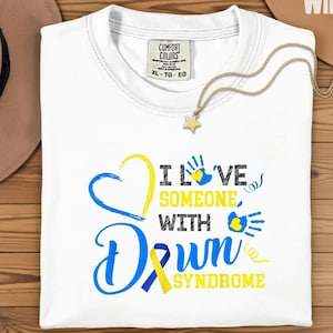 May include: White t-shirt with the text "I LOVE SOMEONE WITH DOWN SYNDROME" in blue and yellow. The design includes a heart, handprints, and a ribbon. A gold necklace with a star pendant is draped across the shirt.