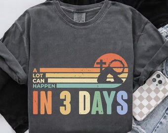 Op de afbeelding: Donkergrijze sweatshirt met de tekst "A LOT CAN HAPPEN IN 3 DAYS" in kleurrijke blokletters. Het ontwerp bevat een kruis, een doornenkroon en een zonsondergang-stijl achtergrond met horizontale strepen.
