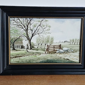 May include: A framed watercolor painting depicting a rural landscape with a large tree, a wooden fence, and a small group of sheep in a field. The artwork is enclosed in a black wooden frame with a thin gold inner border and features a signature.