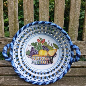 Puede incluir: Una cesta de frutas redonda de cerámica con un diseño azul y blanco. La cesta presenta un borde y asas trenzadas, con una ilustración central de una cesta llena de fruta. La fruta incluye uvas, peras y fresas.