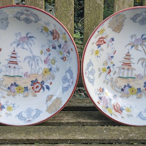 Könnte beinhalten: Zwei weiße Keramikschalen mit roten Rändern und einem Blumendesign mit Pagoden, Palmen und Blumen. Die Schalen stehen auf einer Holzoberfläche.