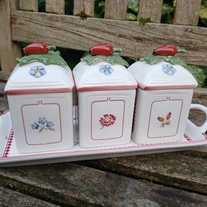 Puede incluir: Tres botes de cerámica blanca con diseños florales y tapas de bayas rojas en una bandeja blanca. Los botes tienen diseños florales de nomeolvides, una flor roja y bayas rojas.