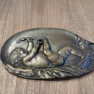 Op de afbeelding: Een ovale bronzen wandplaquette met een reliëfbeeldhouwwerk van een baby die op een doek ligt. De baby houdt een rammelaar in de ene hand en heeft zijn voeten in de lucht.