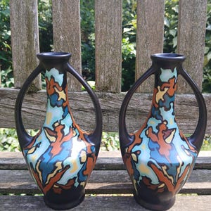 Puede incluir: Dos jarrones decorativos con asas y bases negras. Los jarrones tienen un colorido patrón de formas naranjas, azules y amarillas sobre un fondo blanco. Los jarrones se exhiben en un banco de madera.